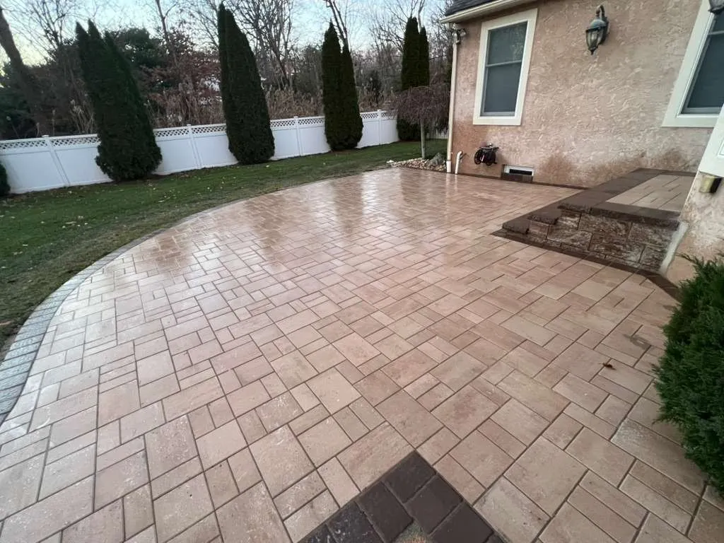 Large tan paver patio with a curved border and stone-faced step behind a stucco home